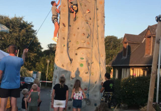 People watch as two climbers scale an outdoor climbing wall at a campsite under a clear blue sky in France.