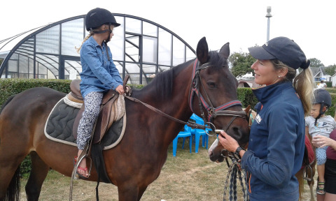 Niña montando un caballo con casco recibe instrucciones de una instructora en Camping Seasonova Etennemare.