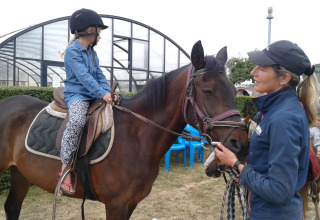 Niña montando un caballo con casco recibe instrucciones de una instructora en Camping Seasonova Etennemare.