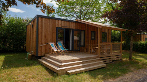 Cabaña de madera con terraza, sillas plegables y rodeada de árboles en un entorno tranquilo y verde.