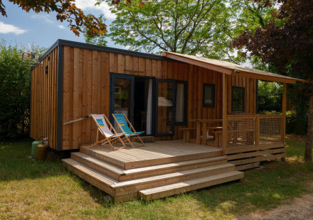 Houten lodge met veranda en ligstoelen, omringd door bomen en groen in een rustige omgeving.
