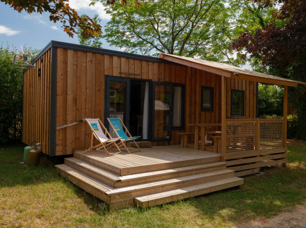 Houten lodge met veranda en ligstoelen, omringd door bomen en groen in een rustige omgeving.