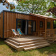 Træhytte med terrasse og liggestole omgivet af grønne træer og buske på en rolig campingplads.