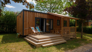 Træhytte med terrasse og liggestole omgivet af grønne træer og buske på en rolig campingplads.