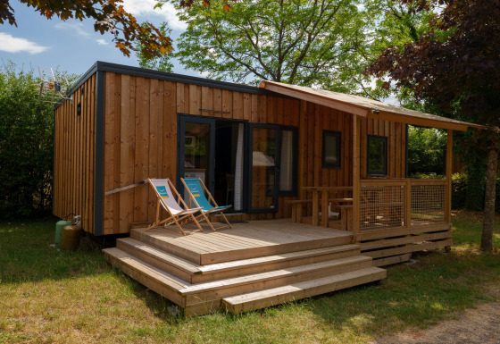 Træhytte med terrasse og liggestole omgivet af grønne træer og buske på en rolig campingplads.