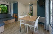 Sala de cabaña moderna con mesa de comedor, sillas blancas, sofá esquinero, TV y ventanas amplias.