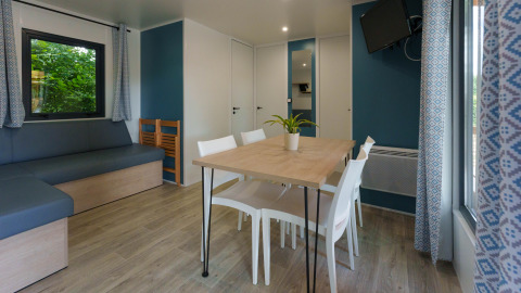 Sala de cabaña moderna con mesa de comedor, sillas blancas, sofá esquinero, TV y ventanas amplias.