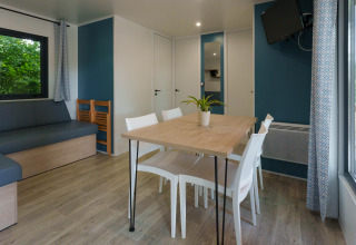 Moderne hytte med spisebord, hvide stole, hjørnesofa, vægmonteret tv og store vinduer med gardiner.