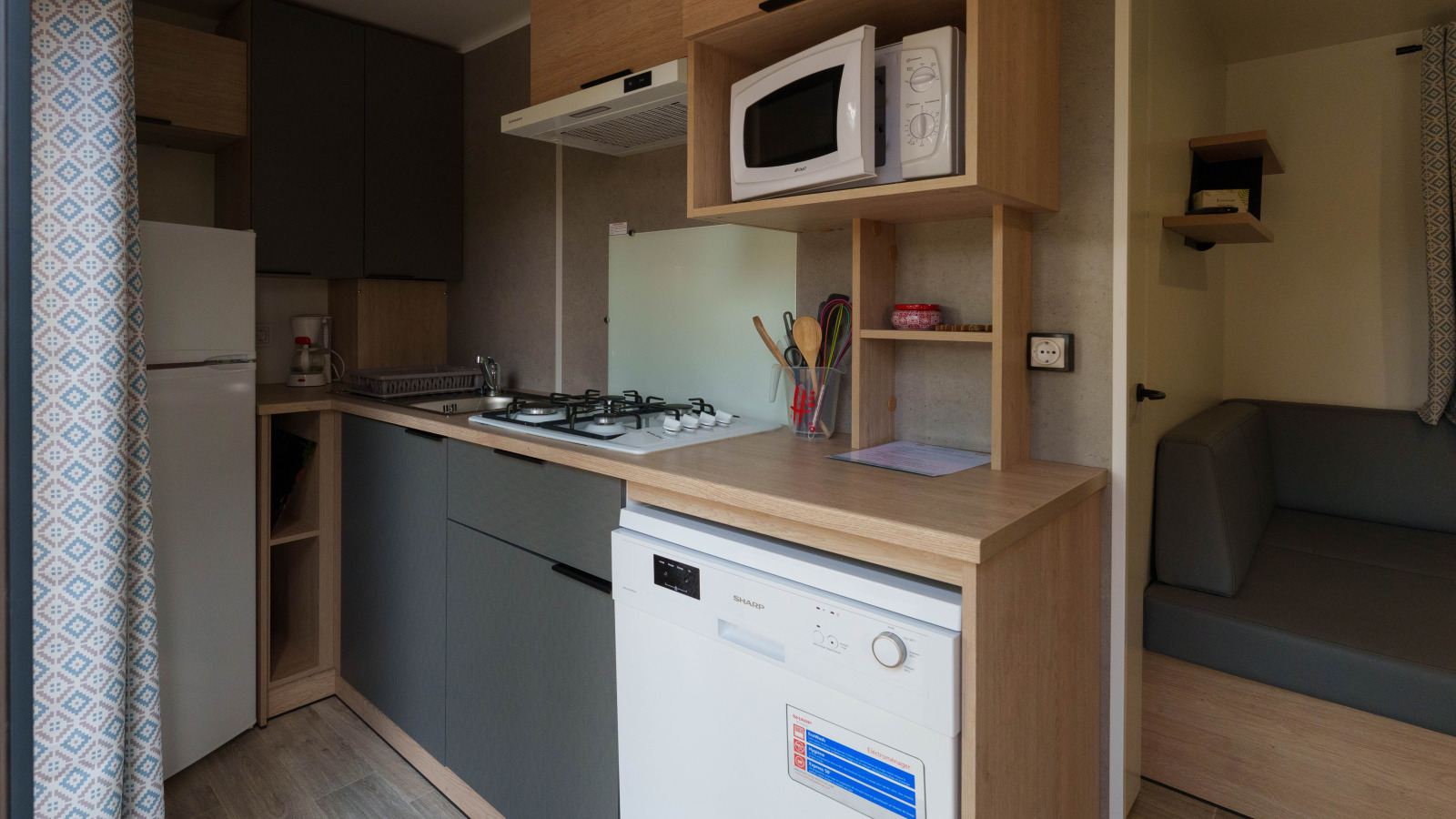 Compacte lodgekeuken met fornuis, magnetron, vaatwasser, koelkast, keukengerei en grijze zithoek.