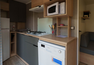 Compact lodge kitchen with stove, microwave, dishwasher, refrigerator, utensils and gray sofa area.