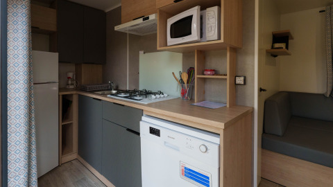 Compact lodge kitchen with stove, microwave, dishwasher, refrigerator, utensils and gray sofa area.