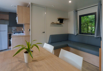 Modern lodge-interieur met eettafel, plantje, hoekzetel, keukentje en raam met gordijnen te zien.