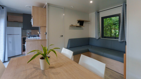 Interior de lodge moderno con mesa de comedor, planta, sofá gris en L, cocina pequeña y ventana con cortinas.