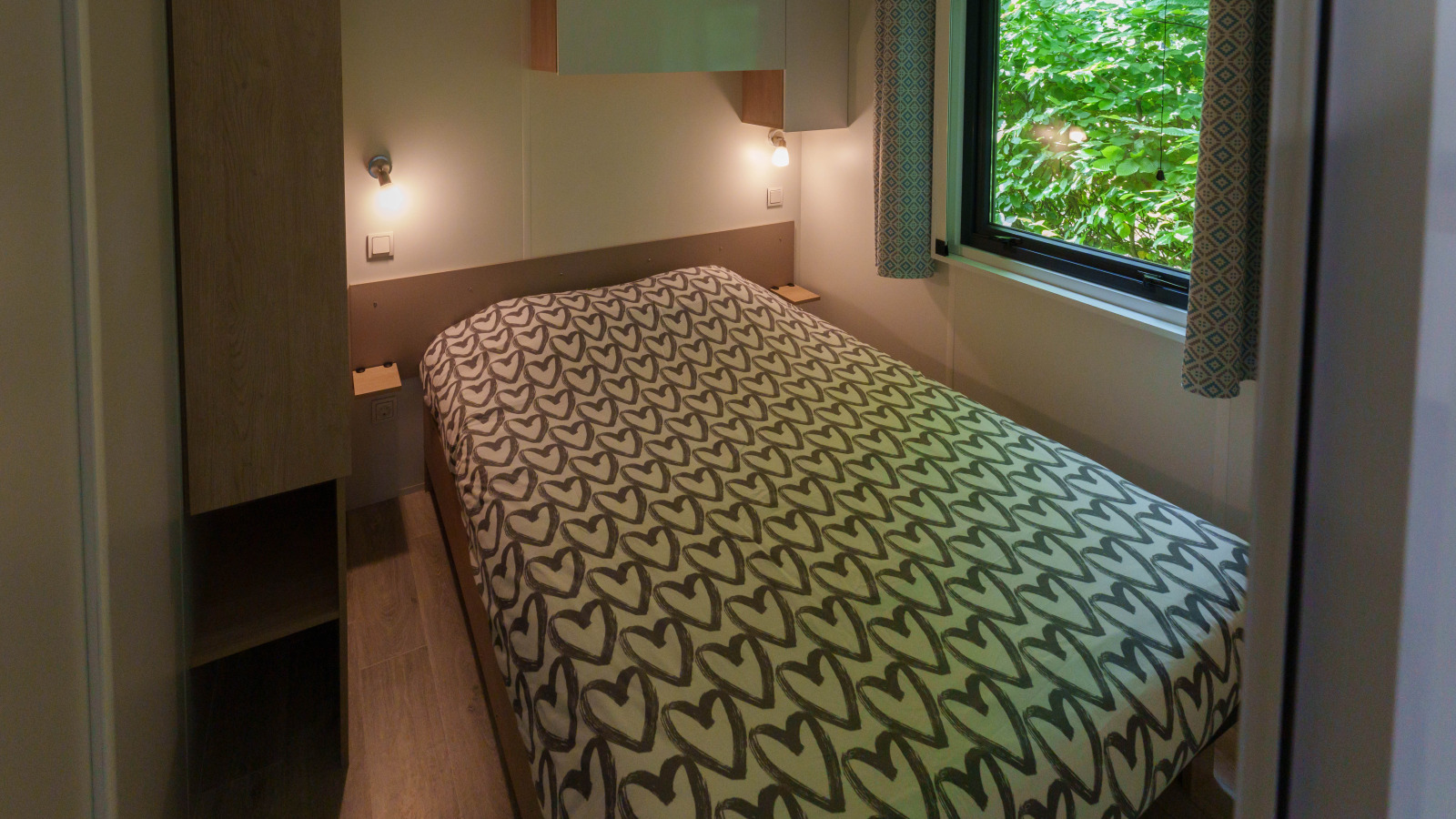 Slaapkamer in lodge met tweepersoonsbed, hartjesdekbedovertrek en raam met uitzicht op het groen.