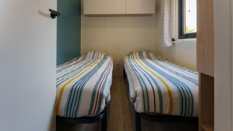A small lodge bedroom with two single beds featuring striped bedding, a window, and wall-mounted cabinets.