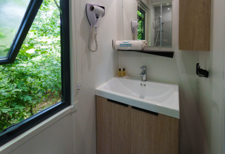 Baño en una cabaña con ventana abierta, lavabo, espejo, secador de pelo y vista a la vegetación exterior.