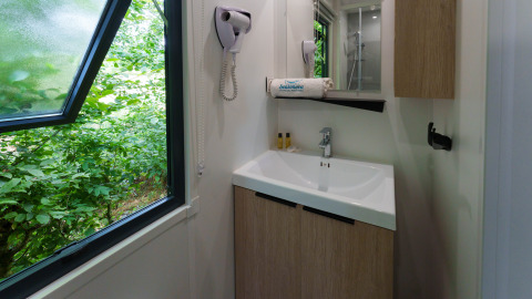 Baño en una cabaña con ventana abierta, lavabo, espejo, secador de pelo y vista a la vegetación exterior.