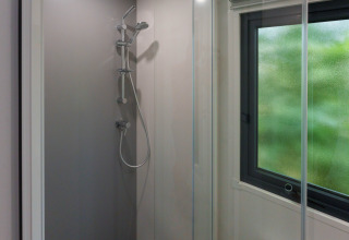 Cabina de ducha moderna con ventana en un lodge, cristal transparente y ducha de mano, luz natural exterior.
