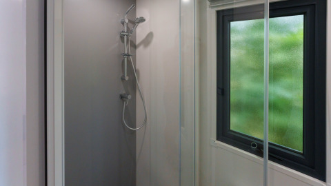 Moderne brusekabine med vindue i en lodge, klart glas og håndholdt bruser, naturligt lys udefra.