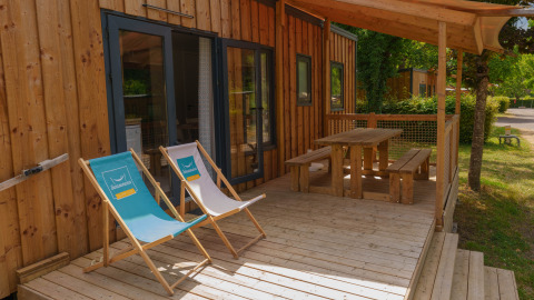 Terraza de madera en una cabaña con dos tumbonas y mesa de picnic bajo toldo, rodeado de naturaleza.