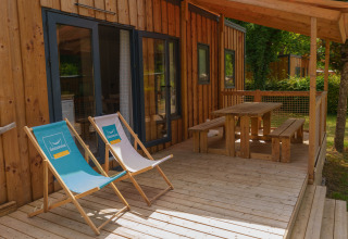 Wooden deck of a lodge with two deck chairs and picnic table under a canopy, surrounded by greenery.