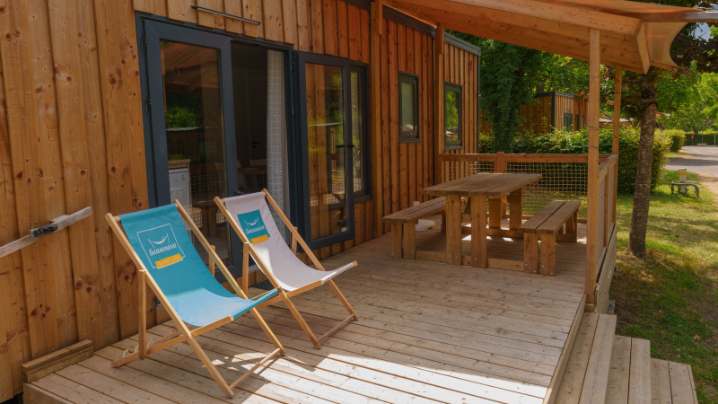 Wooden deck of a lodge with two deck chairs and picnic table under a canopy, surrounded by greenery.