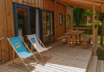 Terrasse en bois d’un lodge avec deux chaises longues et table de pique-nique sous l’auvent, verdure autour.