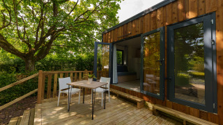 Outdoor deck at a wooden lodge with a table, chairs, and large open glass doors facing the greenery.
