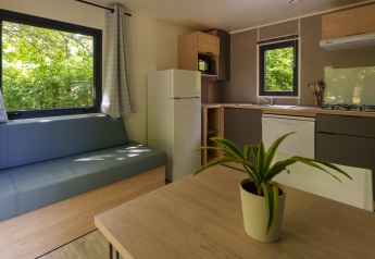 Modern lodge-interieur met eettafel, groene plant, sofa en keuken, met grote ramen en zicht op het groen.