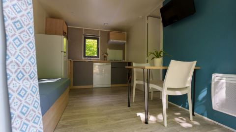 Intérieur de lodge moderne avec cuisine, table à manger, deux chaises blanches, mur bleu et fenêtre.