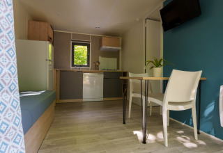 Intérieur de lodge moderne avec cuisine, table à manger, deux chaises blanches, mur bleu et fenêtre.