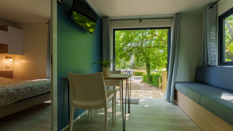 Modern lodge room with bedroom, dining area and open doors leading to a wooden terrace and green trees.