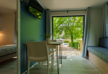 Lodge moderno con camera da letto, zona pranzo e porte aperte su terrazza in legno con vista sugli alberi.