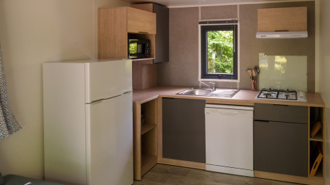 Pequeña cocina moderna en una cabaña con nevera, lavavajillas, estufa de gas y ventana con vista a la naturaleza.