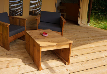 Zona de estar exterior de madera con cojines negros en una tienda safari Slow Lodge en Camping Seasonova Vittel, Francia.