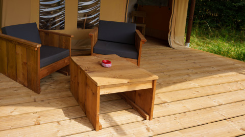 Area salotto esterna con mobili in legno e cuscini neri in una tenda safari Slow Lodge al Camping Seasonova Vittel, Francia.