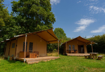 Two safari tents at Slow Lodge beneath a blue sky, surrounded by green trees and grass, with outdoor seating.