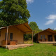 To safari-telte ved Slow Lodge under blå himmel, omgivet af grønne træer og græs, med udendørs siddepladser.