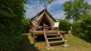 Tenda safari sopraelevata con piattaforma in legno, due sedie e tavolino, circondata da cespugli verdi.