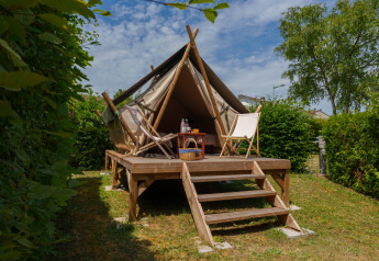 Tienda safari elevada con terraza de madera, dos sillas y mesa, rodeada de arbustos verdes al aire libre.