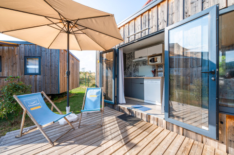 Cottage Prestige lodge deck with umbrella, blue chairs, wooden siding, and open kitchen access in sunlight.