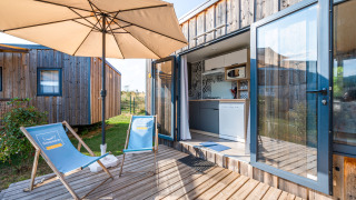 Terraza exterior del Cottage Prestige con sillas azules, sombrilla, deck de madera y vista a la cocina.