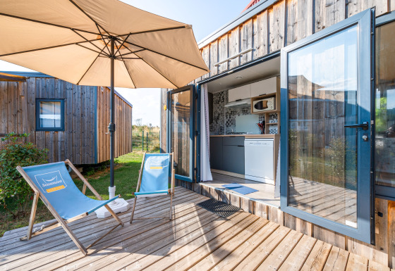 Cottage Prestige lodge deck with umbrella, blue chairs, wooden siding, and open kitchen access in sunlight.