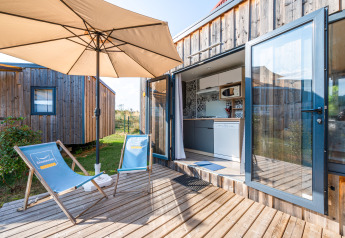 Terraza exterior del Cottage Prestige con sillas azules, sombrilla, deck de madera y vista a la cocina.