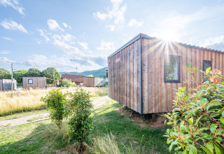 Vista soleada de la cabaña de madera Cottage Prestige en Camping Seasonova Les Portes d'Alsace, Francia, rodeada de naturaleza.