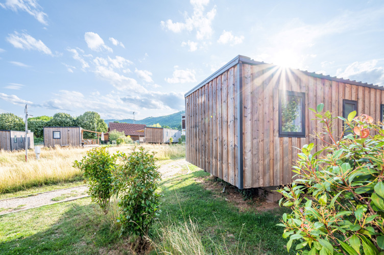 Vista soleada de la cabaña de madera Cottage Prestige en Camping Seasonova Les Portes d'Alsace, Francia, rodeada de naturaleza.