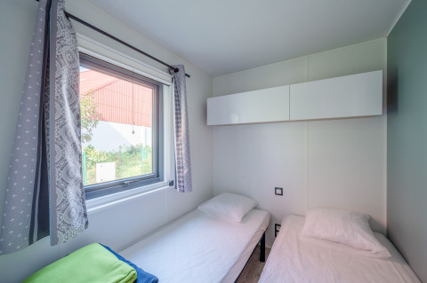 Habitación con dos camas individuales, ventana grande con cortinas y armarios modernos en Camping Seasonova.