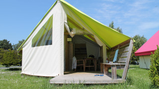 Safari tent met houten terras, bankje en tafel in het gras onder een zonnige blauwe lucht.