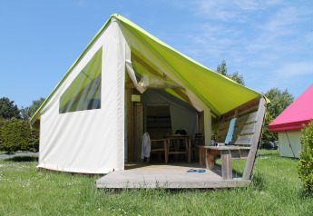 Safari tent with wooden porch, chair and table, surrounded by grass under a clear blue sky.