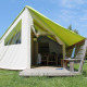 Safari tent with wooden porch, chair and table, surrounded by grass under a clear blue sky.